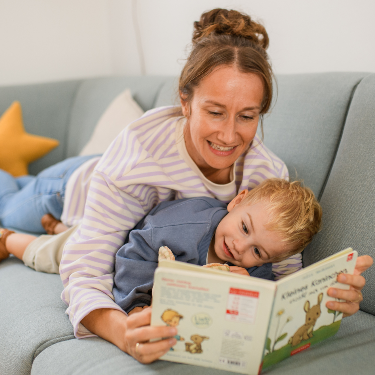 Über uns - Verlage - Vorlesen Foto von Frau mit Kind, die auf dem Sofa liegen und sich zusammen ein Pappbilderbuch anschauen