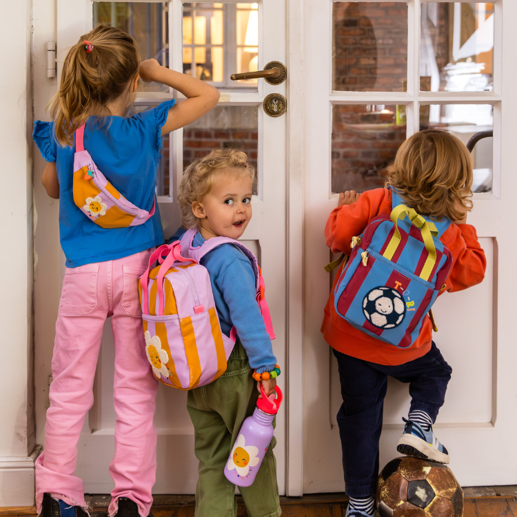 Über uns - Verlage - Spiegelburg Kinder Stimmungsfoto Foto von drei unterschiedlich großen Kindern, die Rucksäcke, Umhängetaschen und eine bunte Trinkflasche tragen und vor einer Glastür stehen, durch die sie teilweise durchschauen
