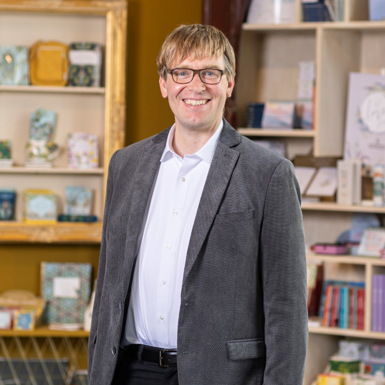 Coppenrath Verlag Geschäftsführung – Dr. Burkhard Scheer Mann mit Brille, weißem Hemd und grauem Sakko vor Regal mit Büchern und Deko