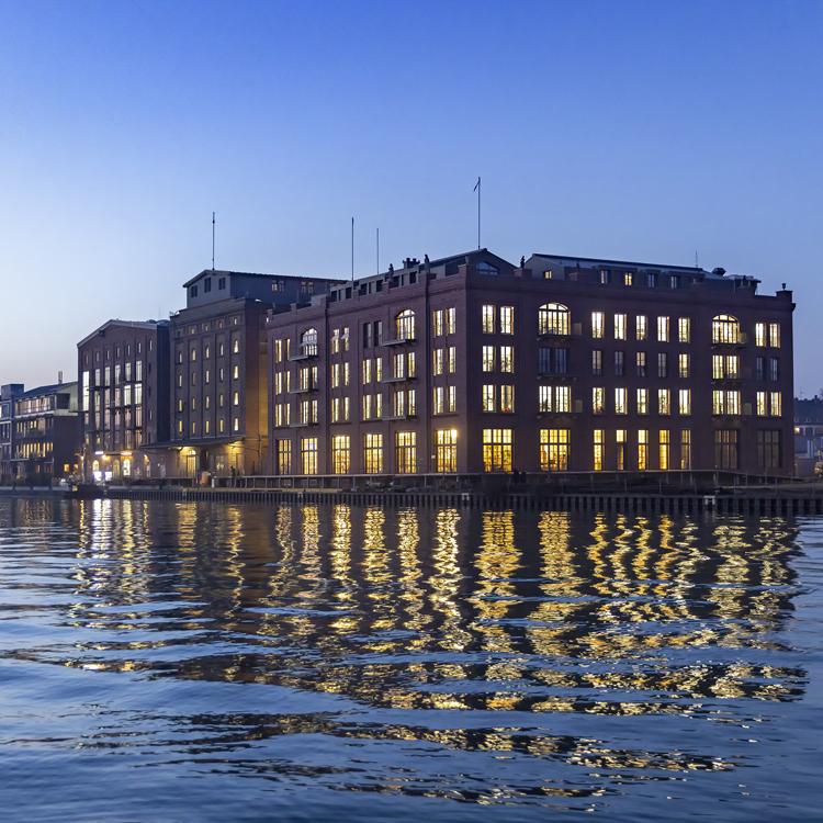 Über uns - Gebäude - Verlag Nacht Kanal Foto im Weitwinkel von mehreren Backsteingebäuden bei Nacht mit leuchtenden Fenstern und reflektierendem Kanalwasser davor