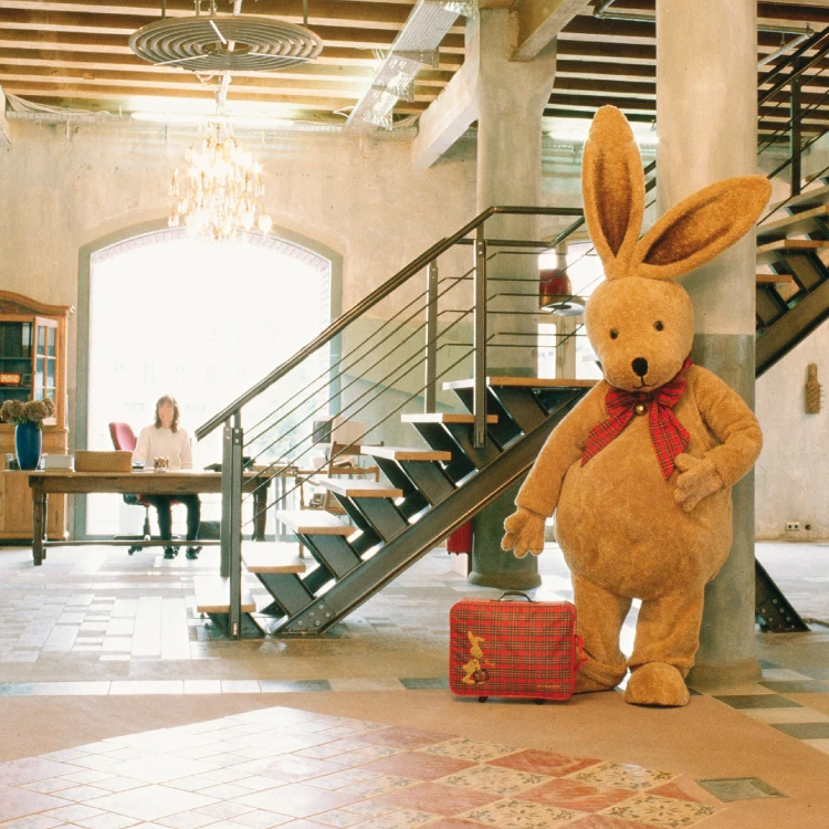 Über uns - Foyer Coppenrath Verlag - Felix der Hase Blick in einen hellen Eingangsbereich mit großer Treppe, einer Frau die an einem Schreibtisch sitzt im Hintergrund und einem großen Stoffhasen mit Koffer im Vordergrund