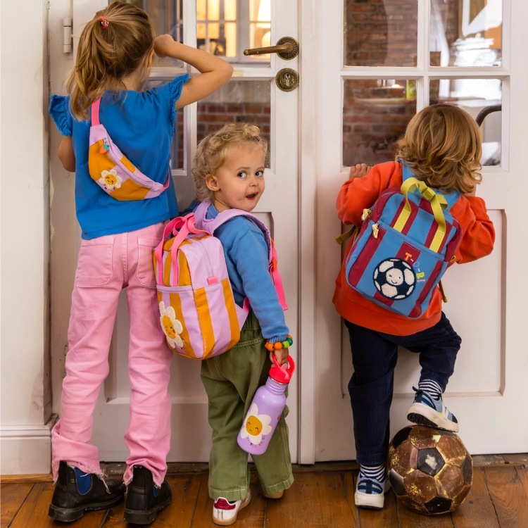 Drei Kinder unterschiedlicher Größer tragen bunte Rucksäcke und Bauchtaschen und schauen durch die Scheibe einer Tür.