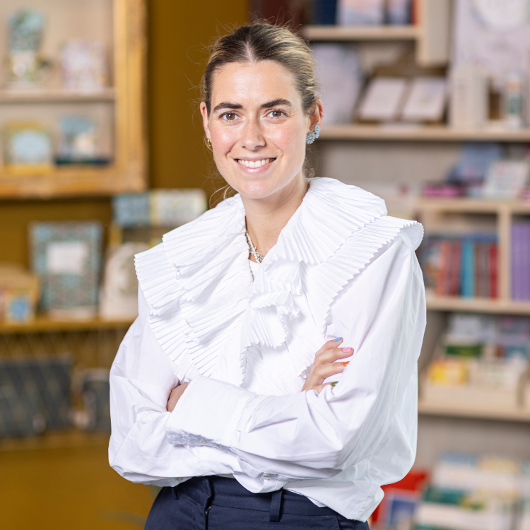 Coppenrath Verlag Geschäftsführung - Louisa Hölker Lächelnde Frau in weißer Bluse mit auffälligem Kragen vor Regal mit Büchern und Deko