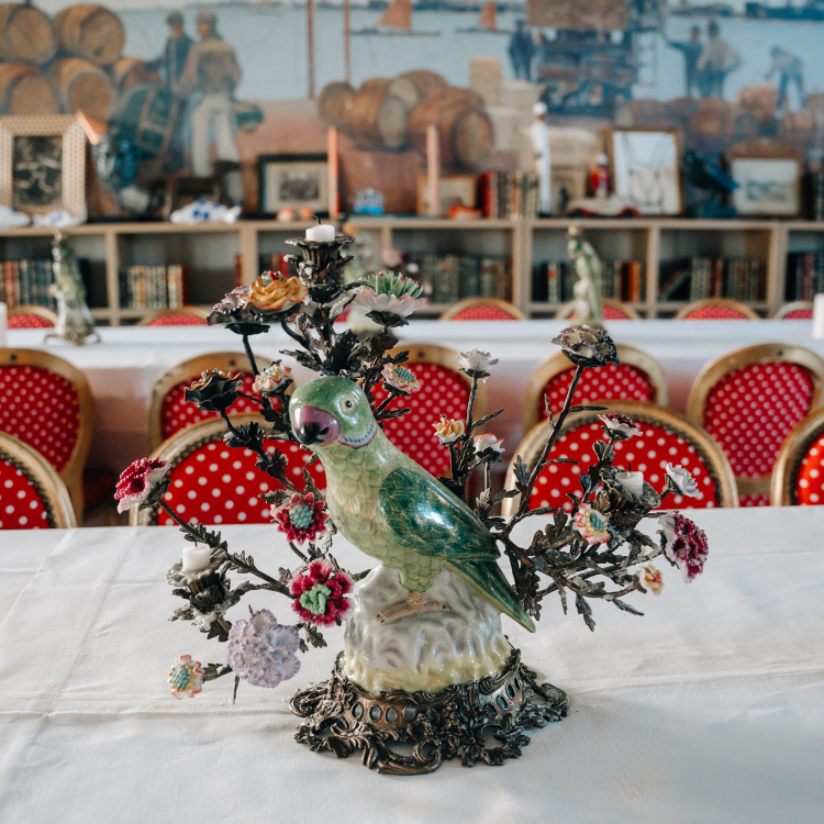 Kerzenhalter mit Vogelstatue Auffälliger Kerzenständer, der die Form eines grünen Papageien hat, steht auf einem Tisch mit weiter Tischtdecke, im Hintergrund rote Stühle mit weißen Punkten