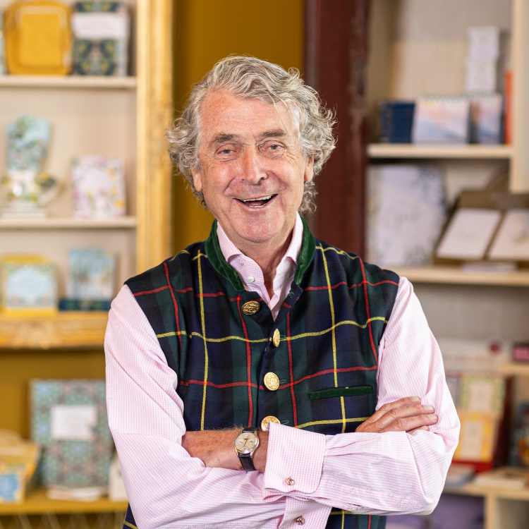 Coppen Verlag Verleger – Wolfgang Hölker Älterer Herr mit grauen Haaren und kariertem Wams steht lächelnd vor gefüllten Bücherregalen