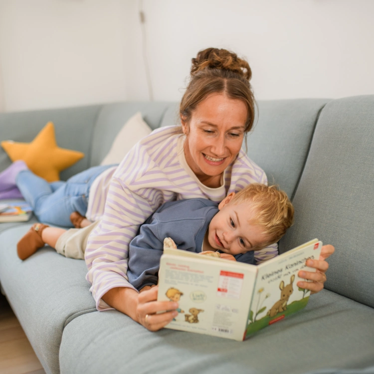Eine Frau und ein kleiner Jungen liegen gemeinsam auf einem Sofa und schauen ein Bilderbuch an.