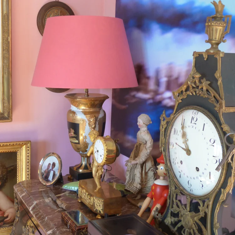 Foto von vielen Vintage Dekogegenständen, die auf einer alten Truhe stehen: eine goldene Uhr, Büsten und eine alte Tischlampe mit pinkem Lampenschirm und geoldenem Ständer